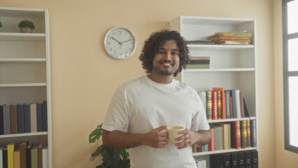Man holding a coffee cup and sipping with both hands in a building office near bookshelves and a wall clock  contentment. © Krakenimages.com