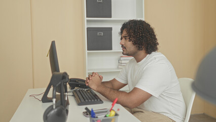 Man at computer covering mouth with hands and clasping at desk beside monitor and keyboard in building with bookshelves visible  relief. © Krakenimages.com