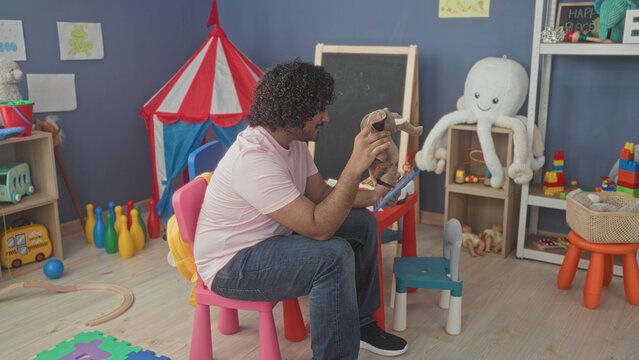 Man holds tablet and stuffed elephant puppet while seated in a kindergarten playroom inside a school building; playful care.