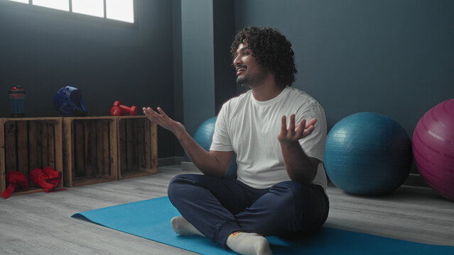 Man seated crosslegged on blue mat gesturing with hands near exercise balls and wooden crates in studio with window light; calm reflection.