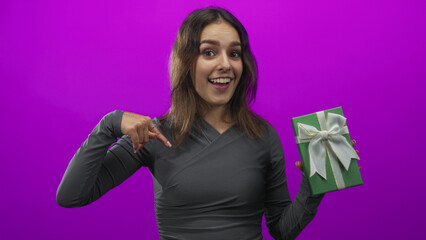 Naklejka premium Woman pointing finger to green gift box while holding present in magenta studio; celebration surprise joy.