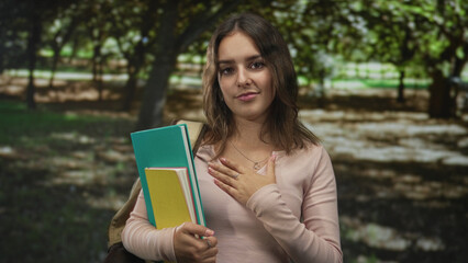 Fototapeta premium Woman holding notebooks and folders, hand on chest and backpack strap visible standing under trees in park; gratitude.