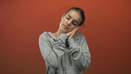 Woman resting head on hands with closed eyes in gray hoodie against orange studio wall and relaxed posture, palms together under cheek  calm. © Krakenimages.com