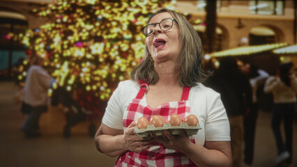 Naklejka premium Woman holding a carton of brown eggs with both hands, smiling while wearing an apron and glasses in building; holiday joy baking.