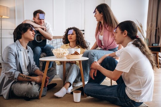 Group of five young adults playing a guessing game with sticky notes on their foreheads, sitting on a rug in a cozy living room with snacks and a guitar