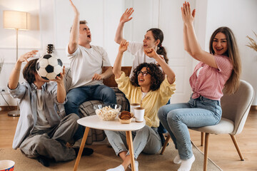 Group of young adults celebrating and cheering while watching a soccer game at home, with snacks and drinks on a coffee table in a bright living room © standret