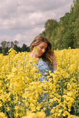 Fototapeta premium A beautiful pregnant woman with long wavy hair, wearing a light blue floral dress, gently holding her belly while standing in a blooming yellow rapeseed field. 