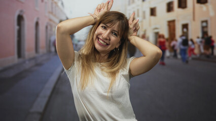 Woman smiling and making bunny ears with hands on a street in the city, wearing a white tshirt and hoop earrings  playfulness. © Krakenimages.com