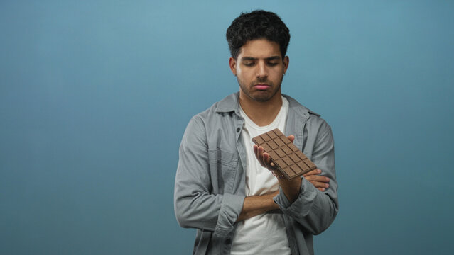 Man holding chocolate bar with one hand, arms crossed and inspecting unwrapped squares against a blue backdrop in a studio; quiet contemplation.