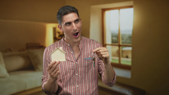 Man holding a wooden house model and keys with open mouth in a living room building; homeownership pride.