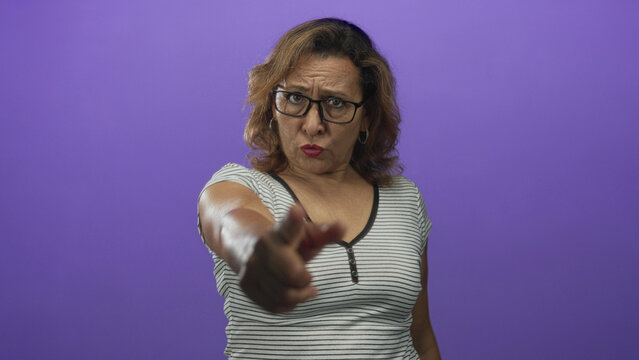 Middle aged hispanic woman pointing finger forward in purple studio; assertion invitation seriousness.