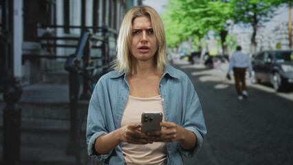 Fototapeta premium Woman holding smartphone with hands, typing and frowning while looking at the screen on a street in the city; concern.