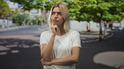 Fototapeta premium Young blonde woman touching chin with finger in street wearing white tshirt and squinting; pensive decision.
