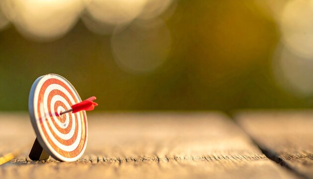 Small red and white target with concentric circles on wooden surface, arrow precisely hitting center, softly blurred warm sunlight background