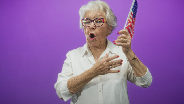 Elderly woman with white hair holding american flag, hand to chest and mouth open singing pledge in studio; patriotic pride.