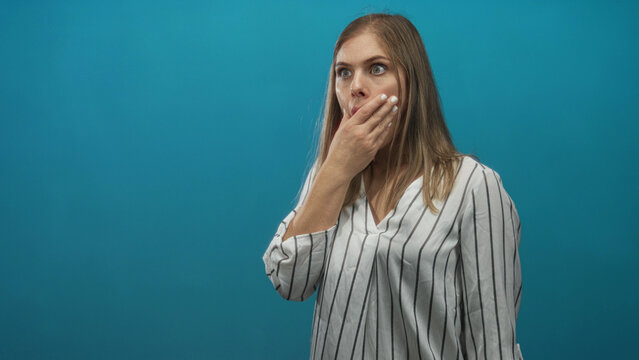 Young blonde woman with finger to lips for silence in blue studio with surprised wide eyed gaze; shock.