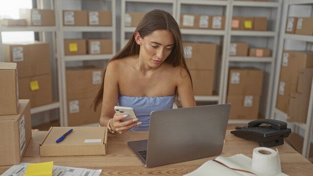 Woman with bare shoulders holding smartphone and checking parcels at a packing desk in a building office; small business determination.