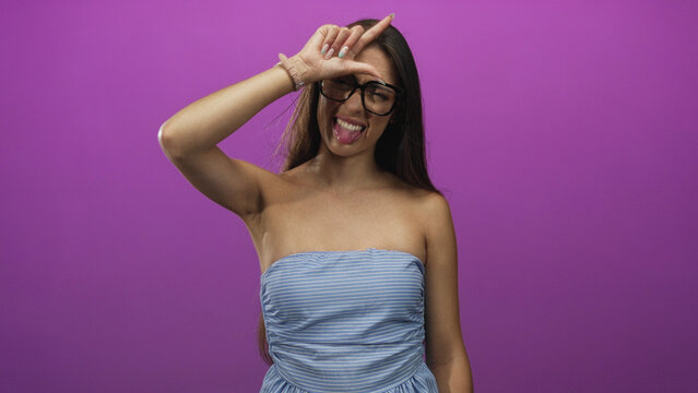 Woman sticking tongue out and making l sign on forehead in purple studio wearing strapless blue dress; playful young fun.