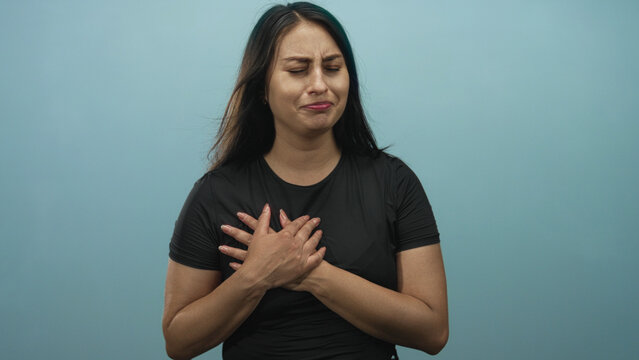 Young hispanic woman clutching chest with both hands crossed over upper chest, eyes closed, wearing black shirt in blue studio; pain.