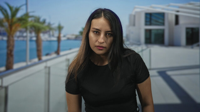 Woman face looking at camera on seaside promenade with glass railing and palms; quiet reflection serenity.