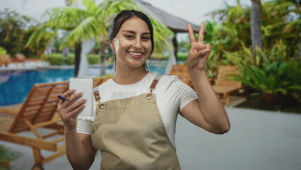 Woman waitress wearing apron and smiling, holding order pad and flashing peace sign at resort poolside  welcoming service. © Krakenimages.com