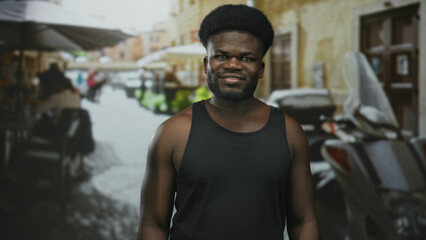 Obraz premium Young man with bare shoulders smiling and standing on a street terrace near scooters and cafe tables; confidence.