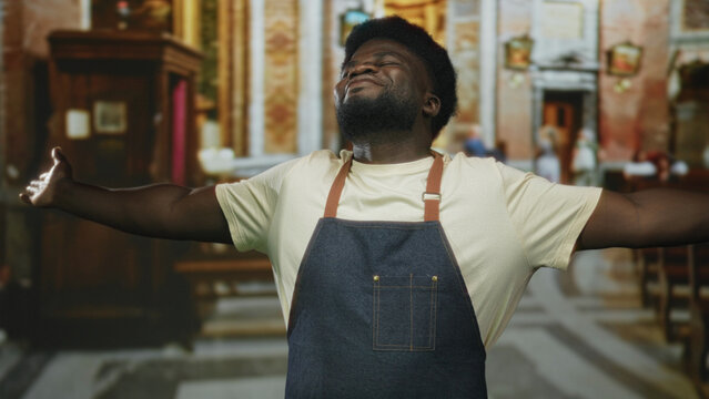Man with arms outstretched in church, wearing denim apron and cream t shirt, head tilted upward and eyes closed; faith gratitude joy.