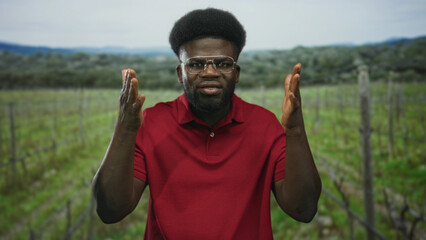 Naklejka premium Man with hands held apart in a vineyard field wearing glasses and red polo shirt, afro haircut and beard; thoughtful reflection.