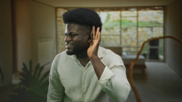 Man cupping ear with hand while listening in a living room building by large window and indoor plant; curiosity focus.