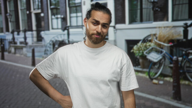 Man with hand on hip and slight smile wearing white t shirt on a cobblestone street near bicycles and row houses in city; confidence easygoing.