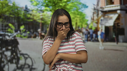 Fototapeta premium Woman with hands on cheeks and glasses on a sunny city street, striped shirt visible; surprise wonder.