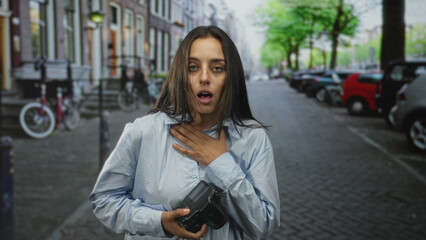 Woman holding camera with hand on chest and open mouth, pointing finger while standing on city street with parked cars and bicycles  surprise. © Krakenimages.com