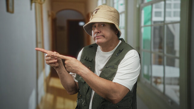 Man fisherman in bucket hat and fishing vest points fingers with both hands and shows a puzzled expression in a building hallway; confusion.
