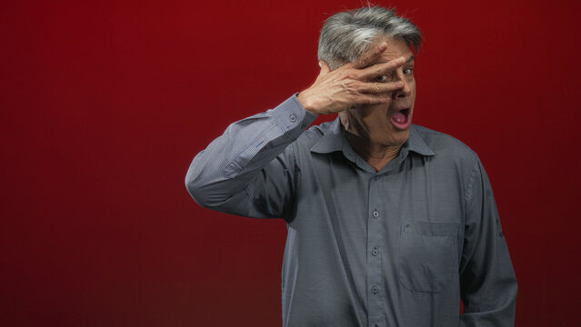 Man with hand covering eye and open mouth gesture in red studio with visible fingers to face and expressive grin; playfulness aging humor.