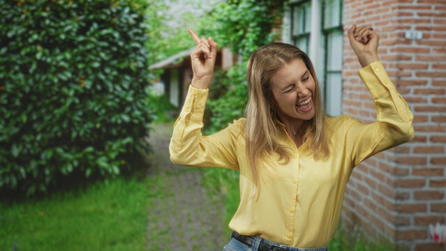 Blonde woman points finger with raised hands near brick building, smiling in yellow shirt and jeans; joy celebration.