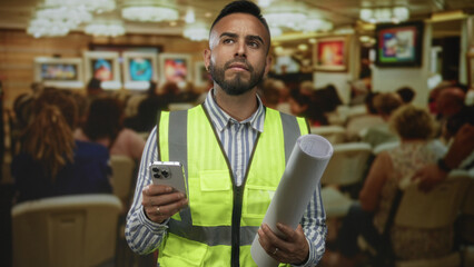 Man holding rolled plans and smartphone, wearing hi vis safety vest in building  planning determination. © Krakenimages.com