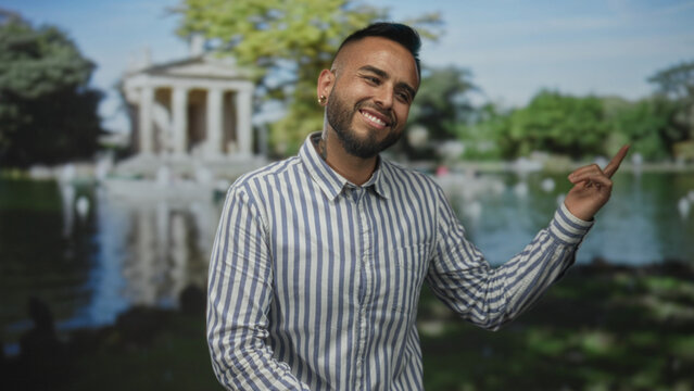 Man smiling with raised finger pointing to building by a park lake, striped shirt visible; joy carefree moment.