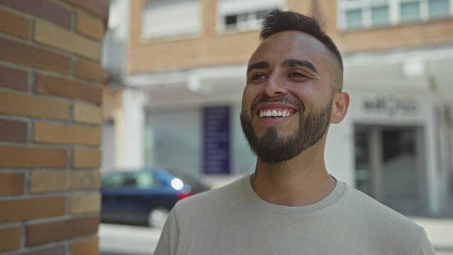 Man smiling with visible teeth and beard on a street next to a brick wall and parked car, facing sideways toward camera; casual happiness.