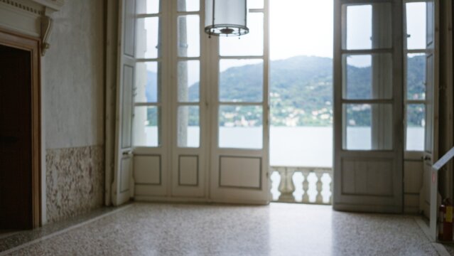 Interior room with open french doors leading to a lakeside balcony, soft defocused bokeh view of water and distant hills; background backdrop copyspace calm.
