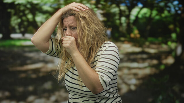 Blonde woman in striped shirt covers mouth with hand coughing in park near green trees and sunlit path, hair loose and windblown; sickness.