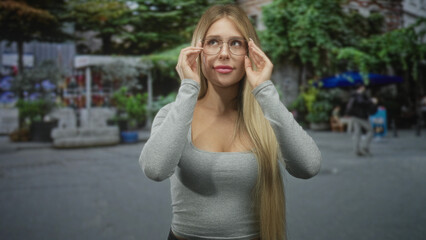 Fototapeta premium Young blonde woman in grey long sleeve shirt adjusts glasses with both hands while looking up in an urban pedestrian street with market stalls; confidence.