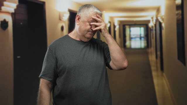 Senior man with hand on forehead and pained grimace, rubbing temple while walking down a building corridor; fatigue aging isolation.