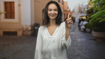 Fototapeta premium Woman smiling with eyes closed pointing index finger while standing on a cobblestone street with potted plants and blurred pedestrians; serenity.