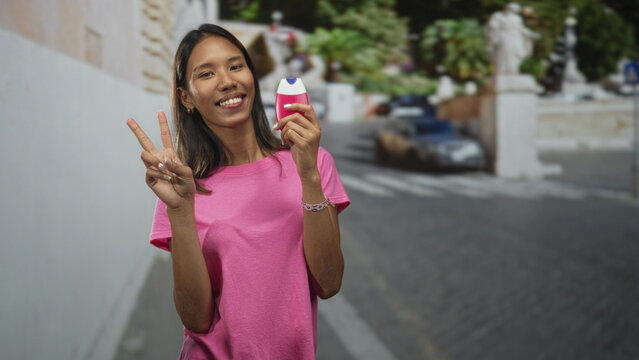 Young thai woman holding a pink toothpaste dispenser, making a finger heart gesture in a street setting; cheerful charm.