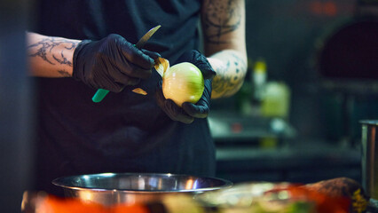 Cook peeling onion with knife over metal bowl in professional kitchen. Concept of restaurant prep, catering service, fresh ingredients, food blog visuals and culinary workflow.
