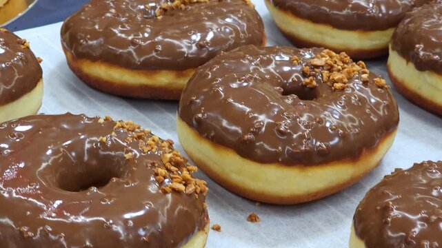 ​Close-up of a row of delicious donuts covered in smooth chocolate frosting and sprinkled with chopped nuts. The pastries are neatly arranged on white baking paper, fresh from the bakery kitchen. 