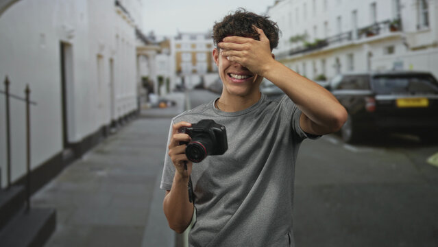 Boy covers eyes with hand while holding dslr camera, smiling with lens cap dangling, wearing gray t shirt on residential street lined with white townhouses and parked cars; shyness playfulness.