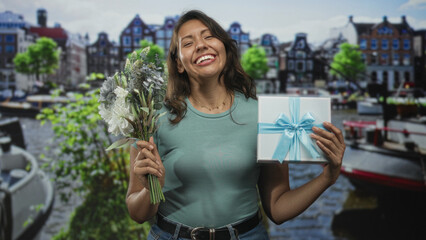Naklejka premium Woman smiling while holding bouquet and gift box on street by amsterdam canal buildings, hands visible and relaxed; joy celebration.