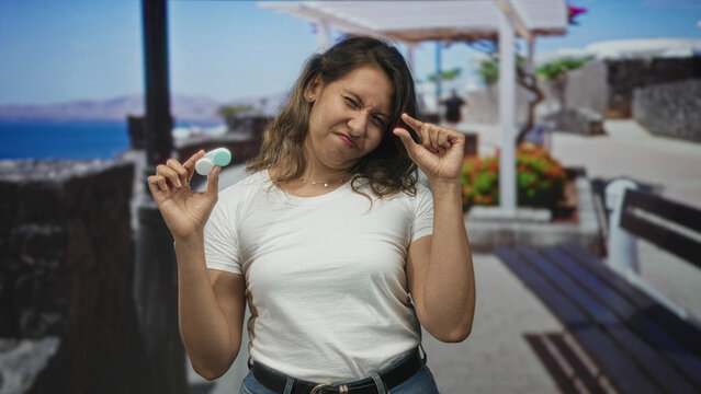 Woman holding contact lens case between fingers on a street promenade near the seaside, pinching tiny lens while squinting in bright sun; disgust.