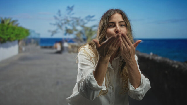 Middle aged hispanic woman blowing a kiss with hand to mouth on a seaside street promenade in a white shirt, smiling and leaning forward; warm greeting affection.